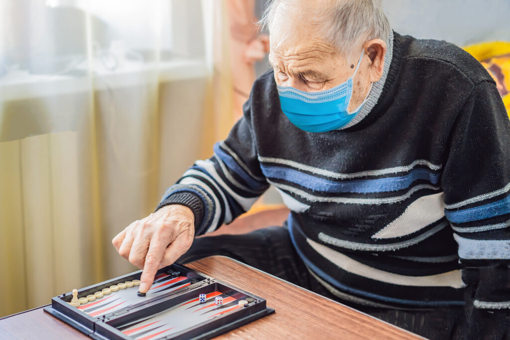Senior Man Wearing A Medical Mask During Covid-19 Coronavirus Pl Senior man wearing a medical mask during COVID-19 coronavirus playing backgammon in a nursing home
