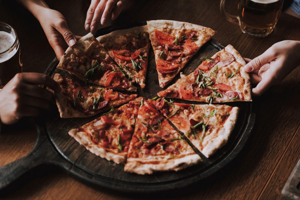 Close Up Of People Hands Taking Slices Of Pizza. Smiling Friends Eating Pizza And Drinking Beer At R