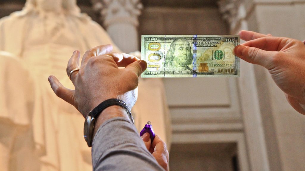  Two new security features on the $100 bill are a holographic 3D ribbon sewn into the paper, and an inkwell that turns into a green bell in the light. (Kimberly Paynter/WHYY) 