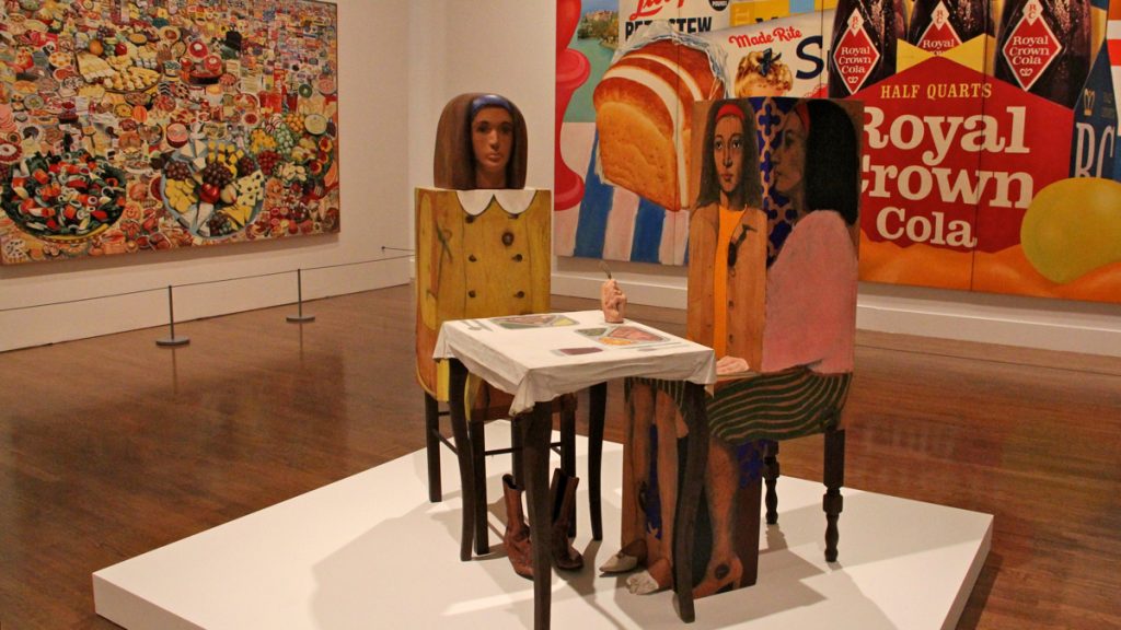 'Dinner Date,'  a sculpture by Marisol, is displayed against the backdrop of Icelandic artist Erro's 'Foodscape' (left) and Tom Wesselmann's 'Still Life #35.'. They are part of the Philadelphia Museum of Art's exhibit on the development of Pop Art. (Emma Lee/WHYY)