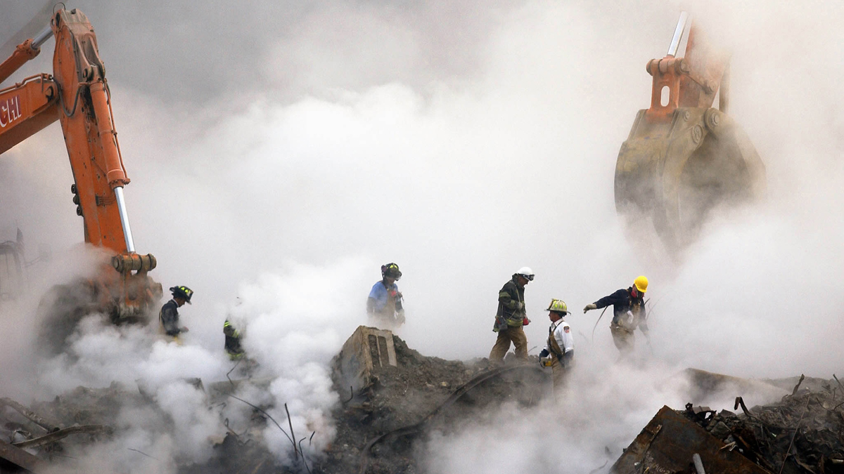 Plumes of smoke rise into the area as firefighters walk over ruins.