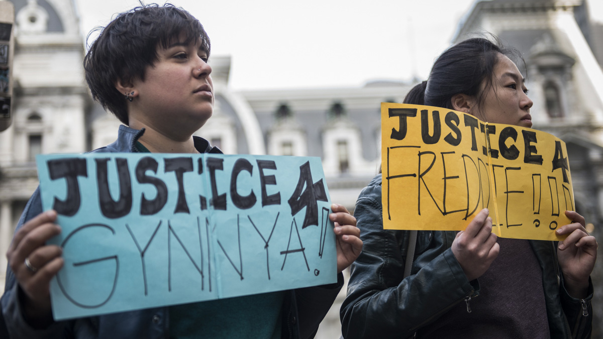 Dimi Xepapadeas (L) and Victoria Yu hold signs seeking justice for Gynnya Mcmillen and Freddie Gray, both of whom were young African Americans that died in police custody.  When asked about what drew her to the case of Freddie Gray, Yu said, “For me it’s very clearly an injustice and needs to be spoken about.” (Branden Eastwood for NewsWorks)