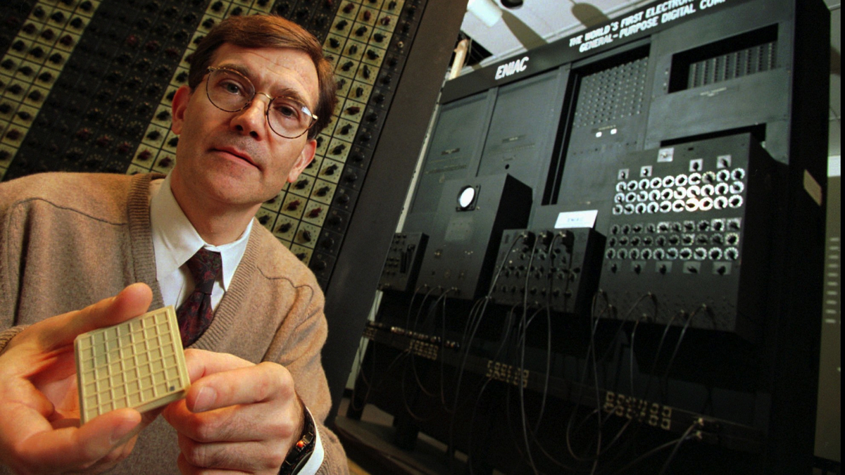 Jan Van der Spiegel holds a microprocessor (the black dot at the lower right corner of the square pad in his hand) in front of about one-tenth of the 50-ton original Electronic Numerical Integrator and Computer (ENIAC)