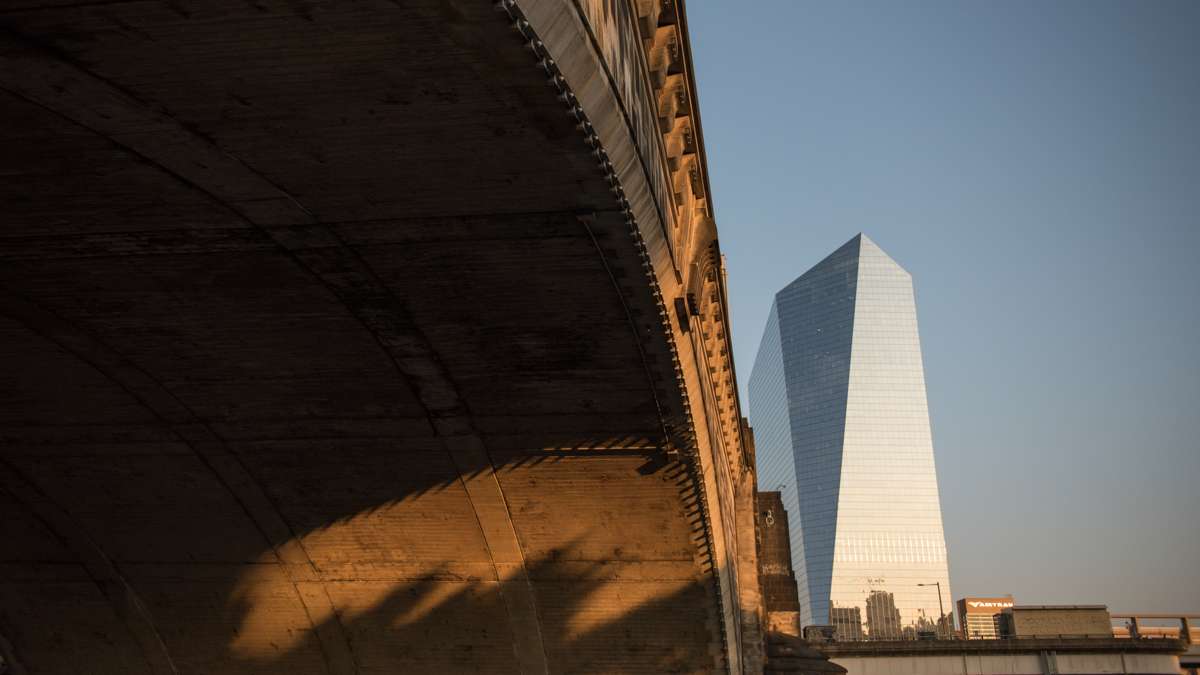 An ornate bridge over the Schuylkill River casts deep shadows while the rising sun illuminates the glass walls of the Cira Center.