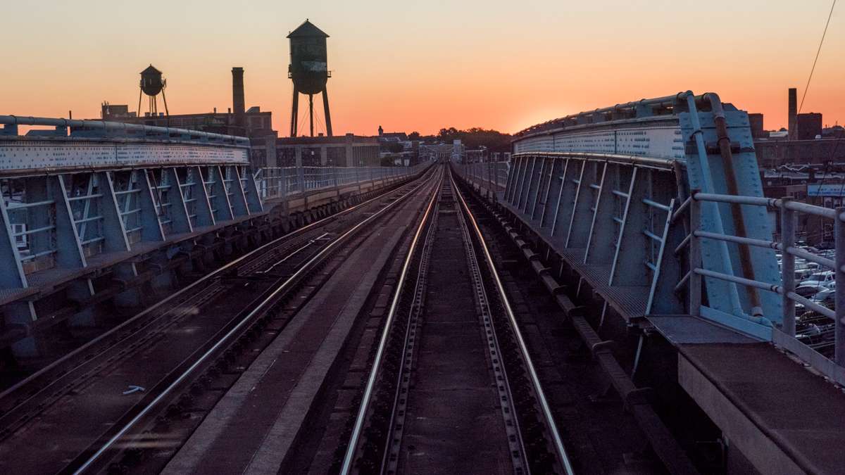 The tracks of the Market Frankford El are cast in purple shadow while water towers and smokestacks are silhouetted against the pale orange glow of the pre-dawn sky.