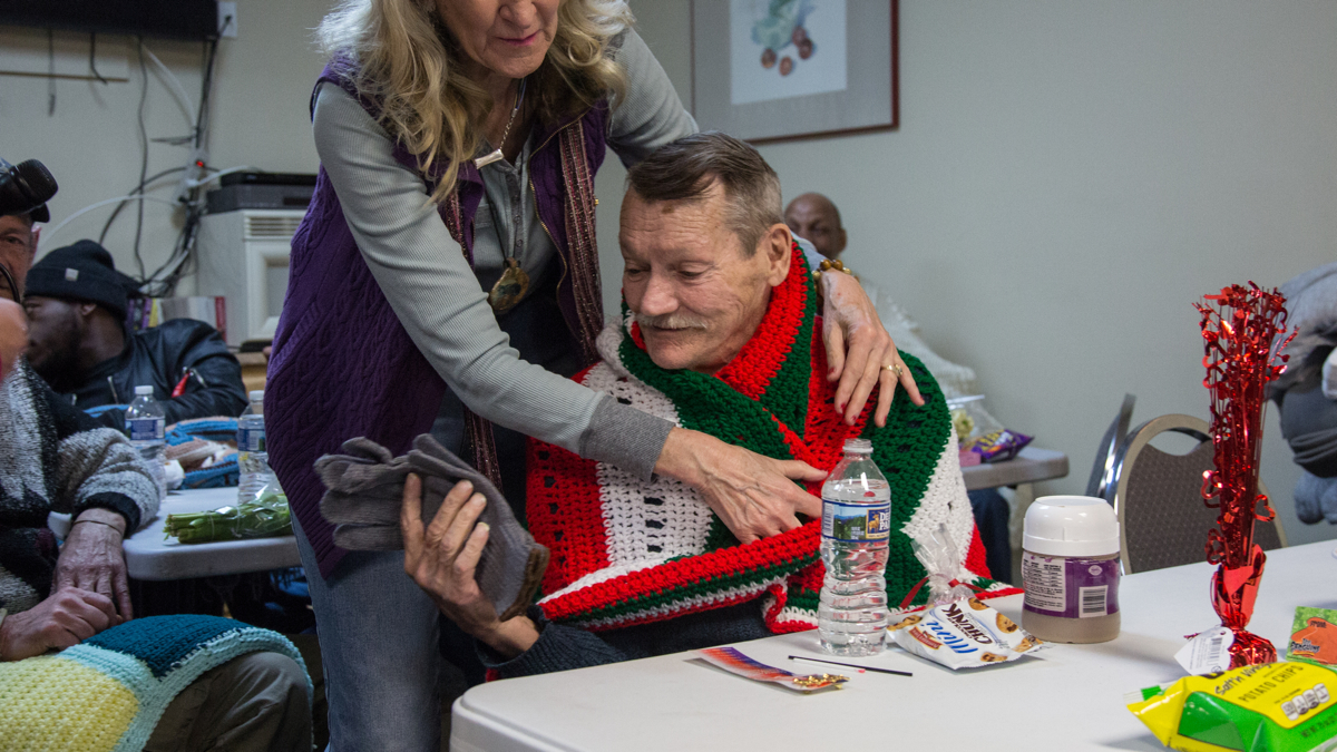 Patricia Gallagher wraps a hand made blanket around the shoulders of a resident at the Open Arms of Nicetown Transitional and Emergency Housing for Vets. Gallager and other volunteers organized a ''Valentine's for Vets'' party and brought vast amounts of donated winter clothing for the men of the shelter. (Emily Cohen for WHYY)