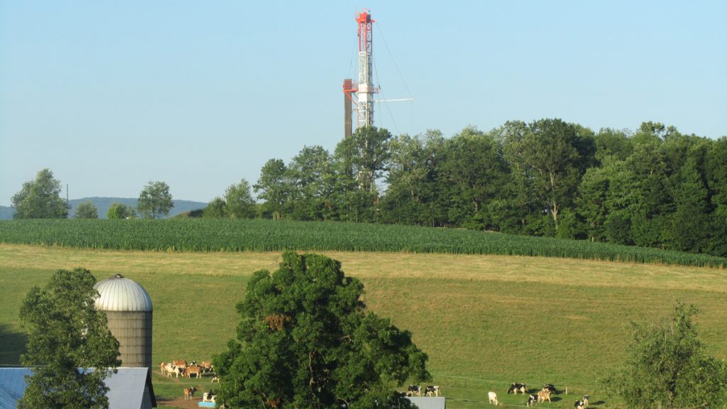NWKPbradfordcountydrillingx1200 A red and white drilling rig justts out in the background behind a line of trees. Farmland and grass, a silo and cows in the foreground