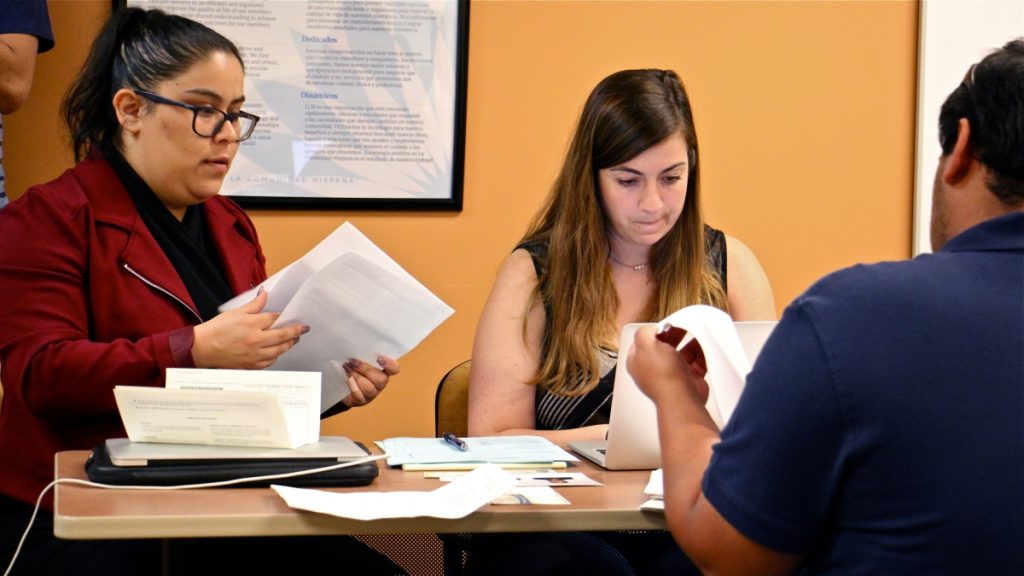 l_img_2354 Two women at a desk facing the camera look over paperwork, a man, on the other side of the desk, his back to the camera