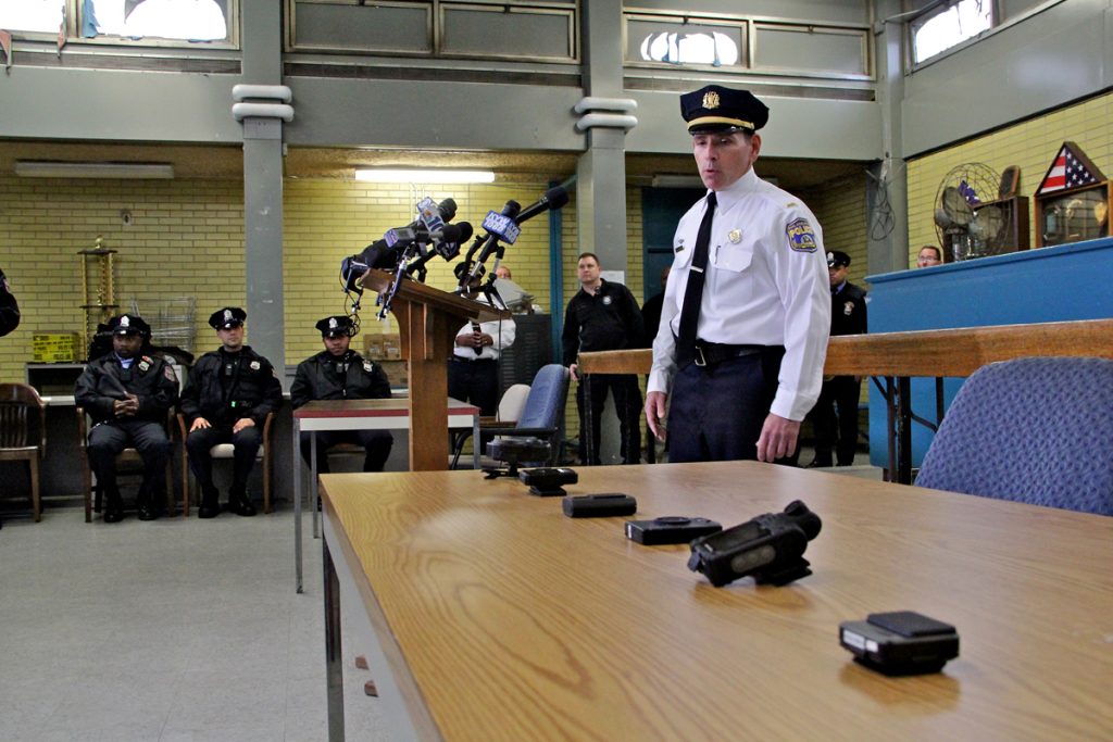 2014 12 11-e lee-tom mclean-philadelphia-police body camera Lt. Tom McLean stands near a table, a half-dozen body camera options are displaued; officers sit and stand in the background