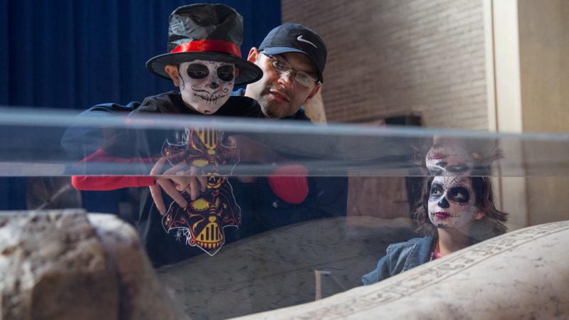 children in skull face paint look at a mummy in a glass case