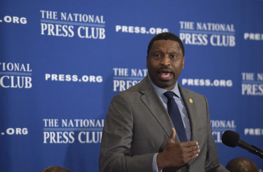 A man in a suit speaks in front of a blue, National Press Club background