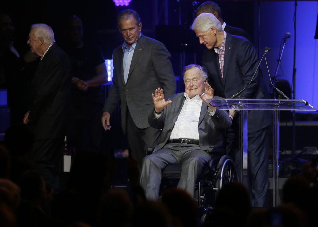 George H.W. Bush,George W. Bush,Jimmy Carter,Bill Clinton President George H.W. Bush in a wheelchair, hands up, palms out, leaving a stage; President Bill Clinton pushes Bush, Sr.; President George W. Bush walks off stage, looking back to his father