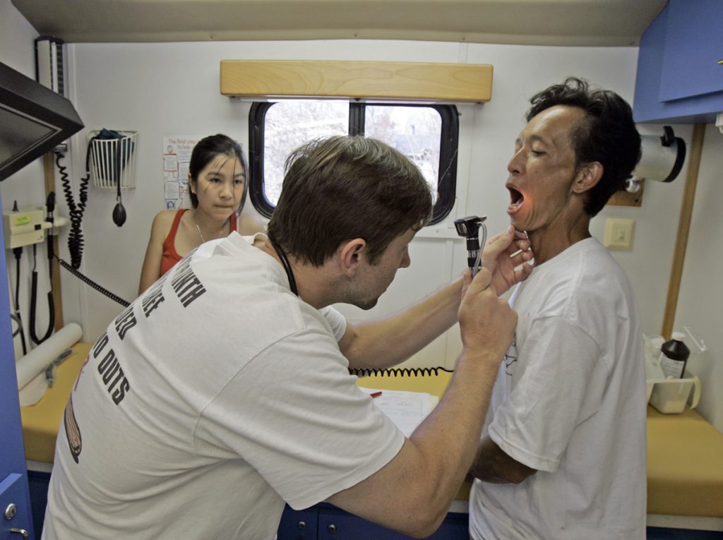 A doctor uses a scope to look into the mouth of a patient; another woman looks on from the rear