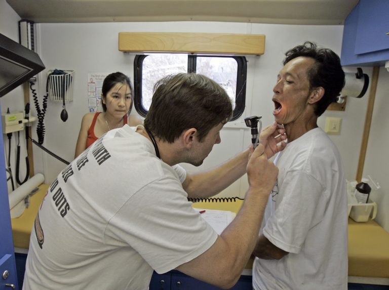 A doctor uses a scope to look into the mouth of a patient; another woman looks on from the rear