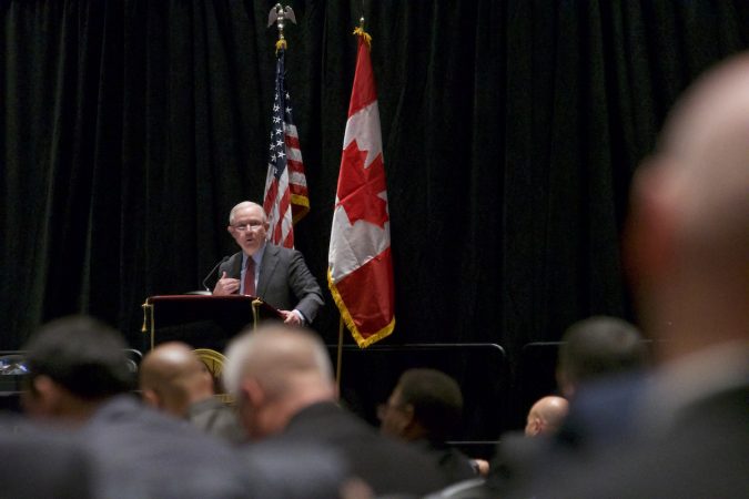 Sessions on stage, the American and Canadian flags stand behind him