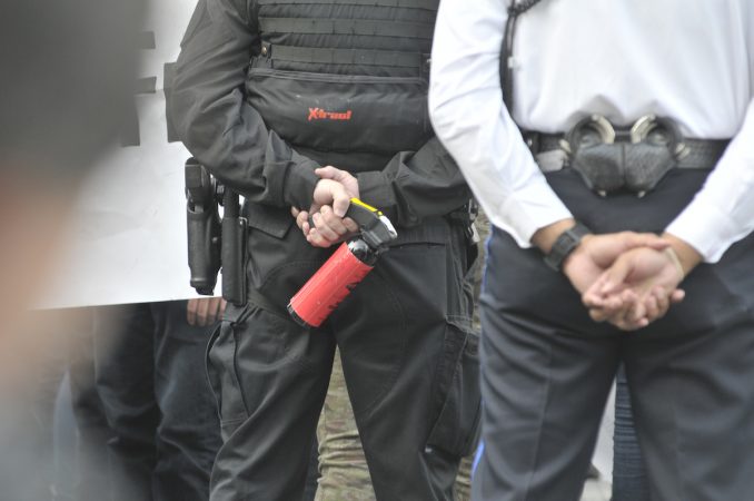 Officers from behind, stand with their arms behind their backs, one holds a can of pepperspray or teargas