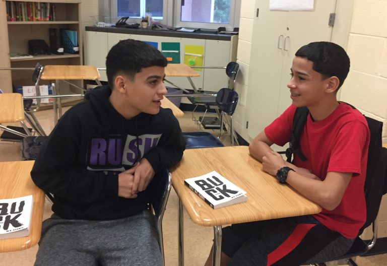 Two schoolboys seated at desks, one turned to the other talking, a book between them, 