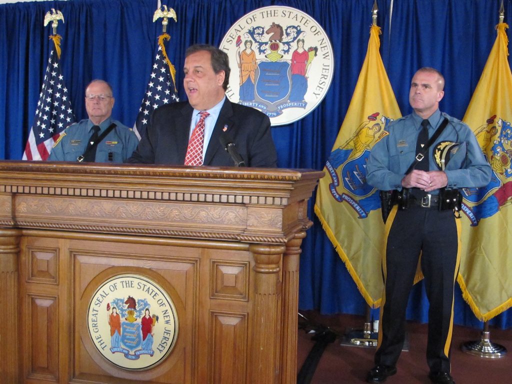 IMG_4030 Governer Christie stands at a podium flanked by two officers, American flag, and New Jersey flag, and seal