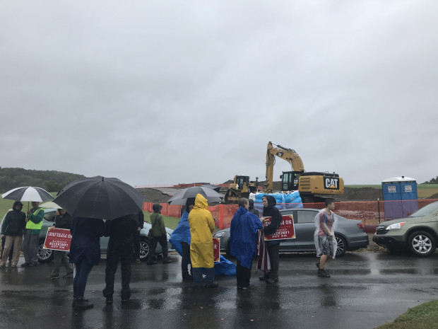 MCSIPAprotestersME2 People stand in the rain, behind them a line of cars, behind that, heavy equipment