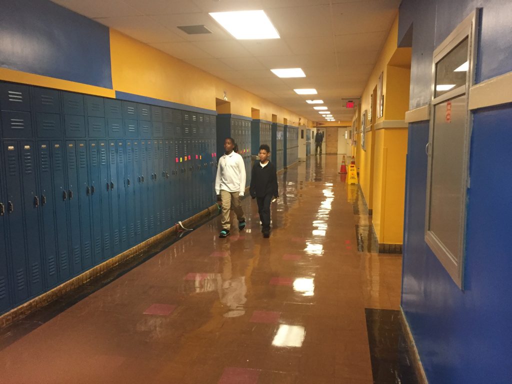 Two students walk towards the camera in an otherwise empty school hallway, blue lockers, yellow walls