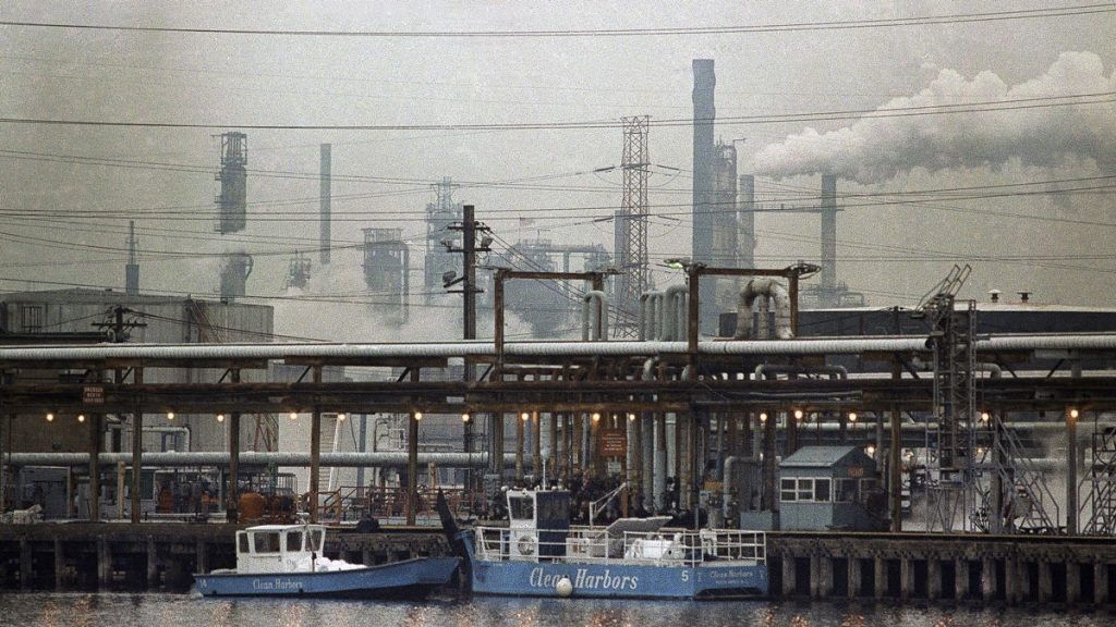 l_bayway two clean harbor cleanup boats work in a waterway alongside an industrial refinery, smoke stacks in the background
