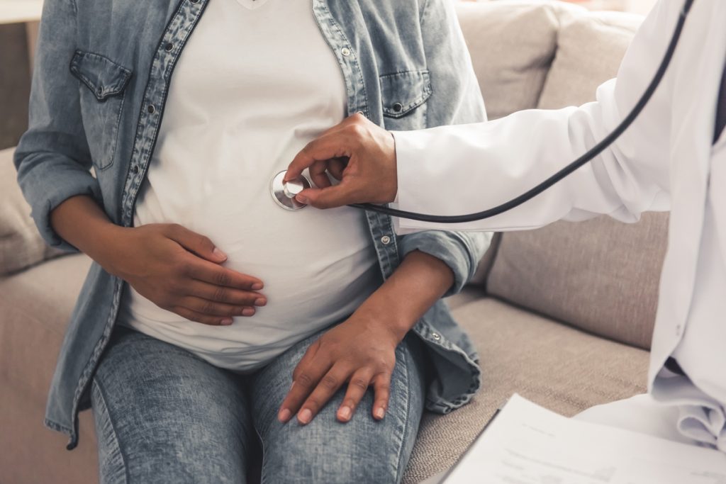 At The Doctor A doctor holds a stethoscope on a pregnant person's belly.