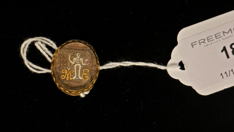 An enameled skeleton holds an hourglass and an arrow in this late 17th century mourning slide. The image is set against intricately woven hair and protected by a faceted rock crystal.