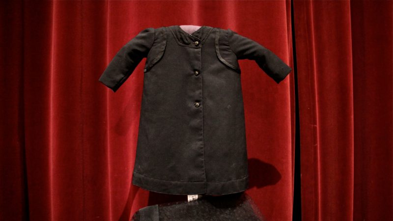 A child's black mourning gown stands out against a red velvet curtain