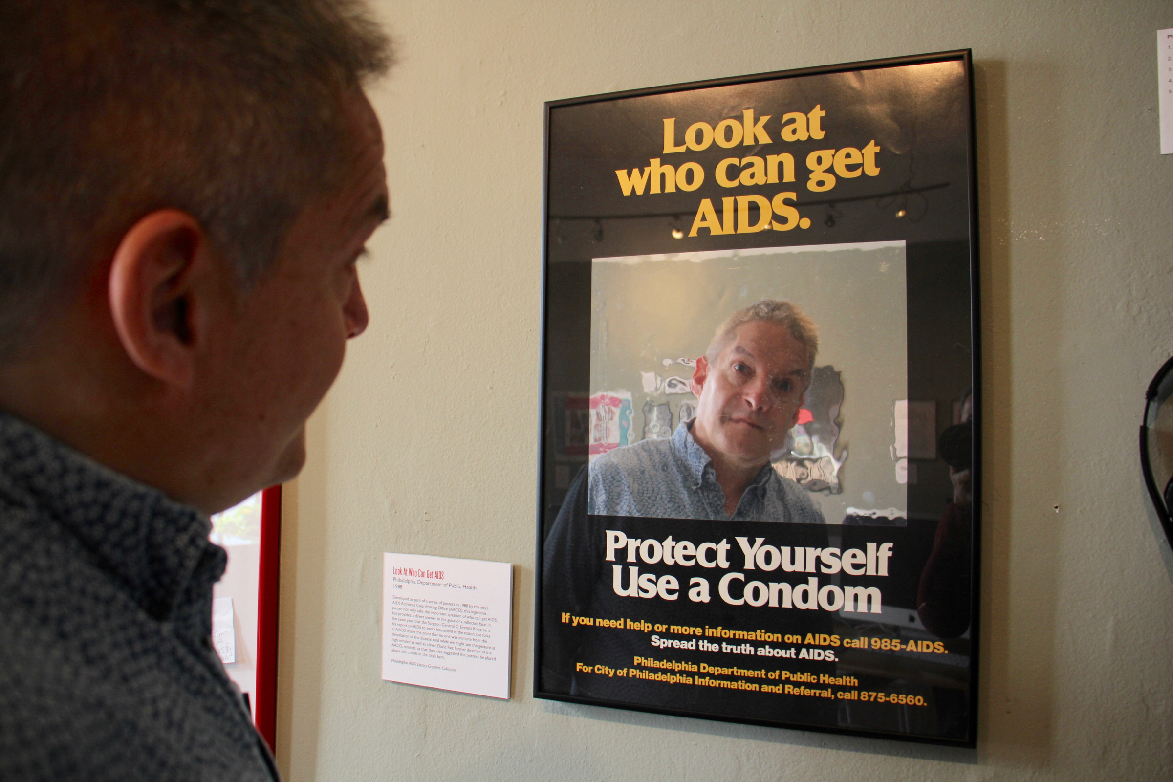 Christopher Bartlett, executive director of the William Way LGBT Center. gazes omtp the reflective surface of a 1988 AIDS awaremess poster.