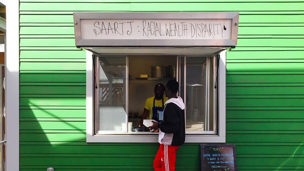 

A customer approaches the window at Saartj, a pop-up food stall in New Orleans running a social experiment. Customers of color are charged the listed $12 price for a meal. White customers are told about the income gap in New Orleans between whites and African-Americans and asked if they want to pay $30 instead, a price that reflects the gap. (Deji Osinulu)