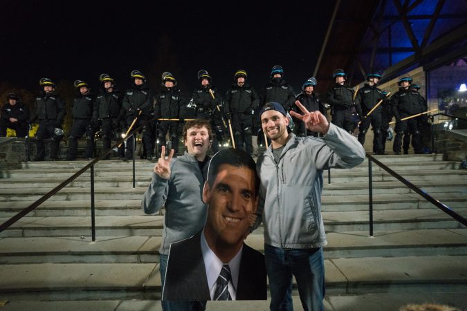 Villanova alums Patrick Frangos, on left, and Mike Arpa pose for a photo in front of riot police with a cut out head of the men's basketball team coach Jay Wright. (Branden Eastwood for WHYY)