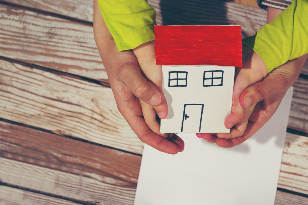 Hands holding paper house on wooden background (jovanmandic/Bigstock)
