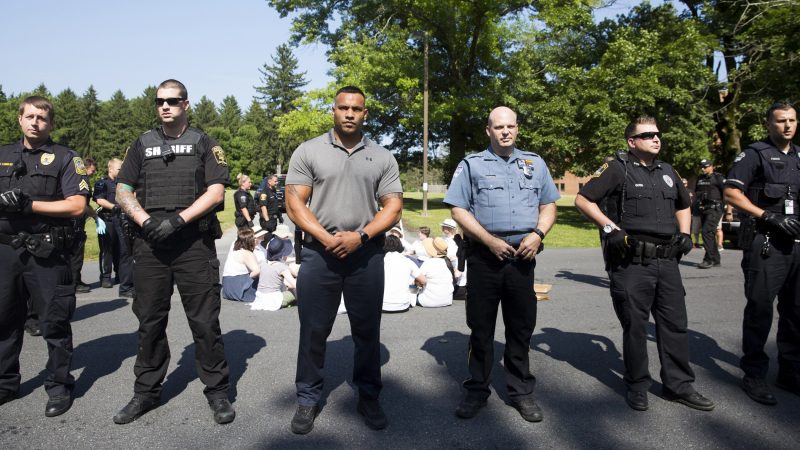 Berks County police officers form a human barricade between seated and standing protestors at the Vigil to Shut Down Berks at the Berks County Residential Center on July 15. (Rachel Wisniewski for WHYY)