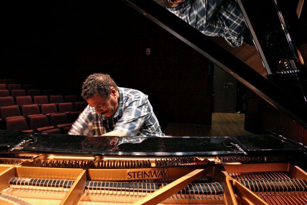 2018 08 01-e lee-alfonso fuentes-princeton university-visiting scholars from puerto rico hurricane piano Composer and educator Alfonso Fuentes plays a black Steinway grand piano under a spotlight in a dark and empty auditorium at Princeton University