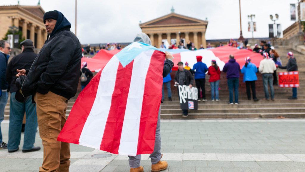 March to Remember the victims of Hurricane Maria A man wears a cape made from a Puerto Rican during an event to remember the people who died in Hurricane Maria in Puerto Rico. The group of activists gathered on the steps of the Philadelphia Museum of Art on Oct. 13, 2018. (Miguel Martinez for WHYY)
