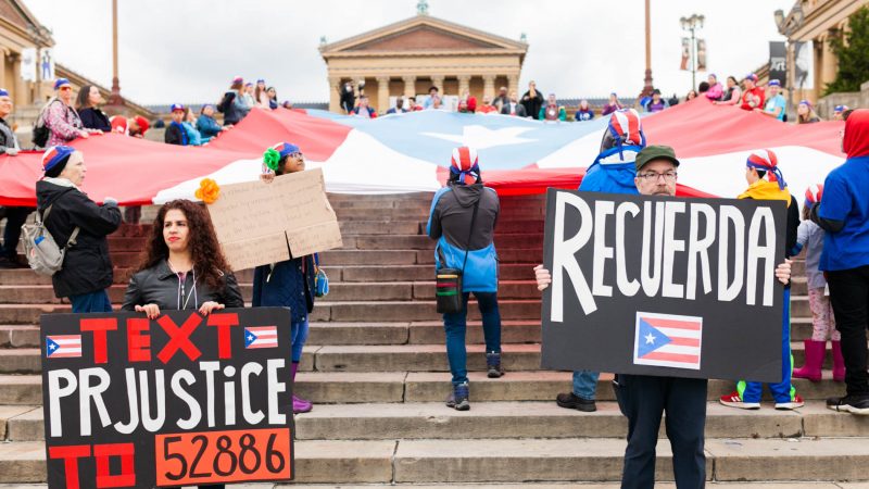 Yecenia Vazquez (left) and Jaime Knowles hold signs in front of a large Puerto Rican flag that was laid it at the steps of the Philadelphia Museum of Art. A group of activist smarched the Benjamin Franklin Parkway to commemorate the people who died in Hurricane Maria in Puerto Rico. (Miguel Martinez for WHYY)