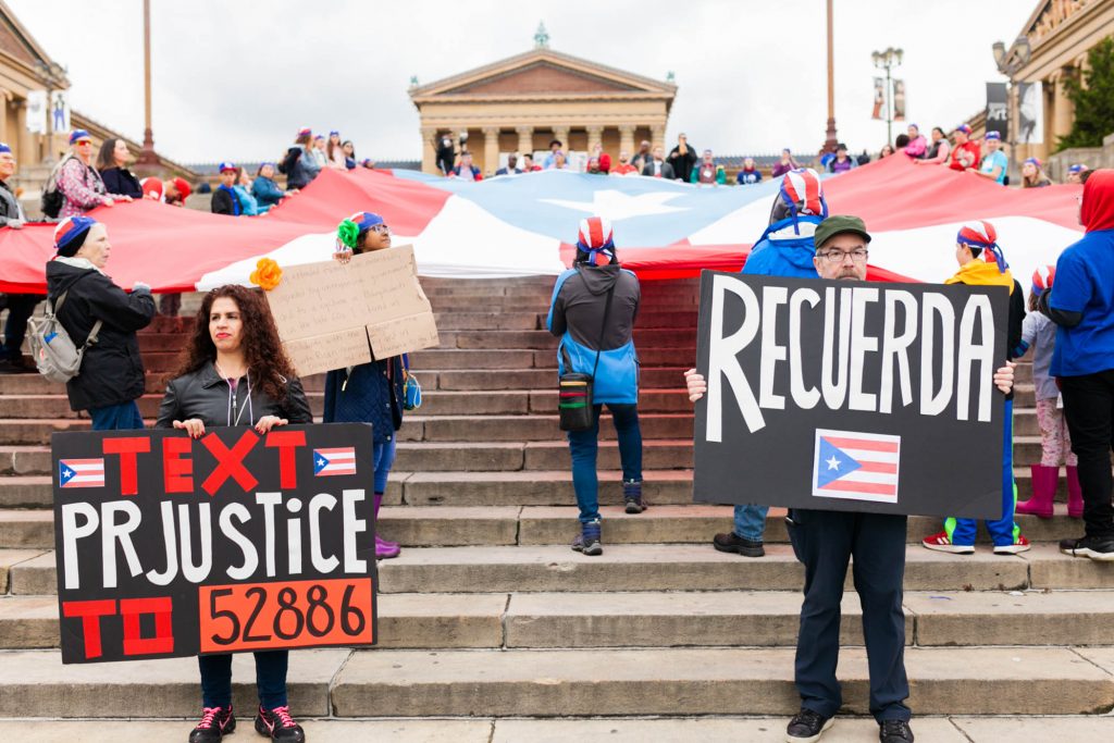 March to Remember the victims of Hurricane Maria Yecenia Vazquez (left) and Jaime Knowles hold signs in front of a large Puerto Rican flag that was laid it at the steps of the Philadelphia Museum of Art. A group of activist smarched the Benjamin Franklin Parkway to commemorate the people who died in Hurricane Maria in Puerto Rico. (Miguel Martinez for WHYY)
