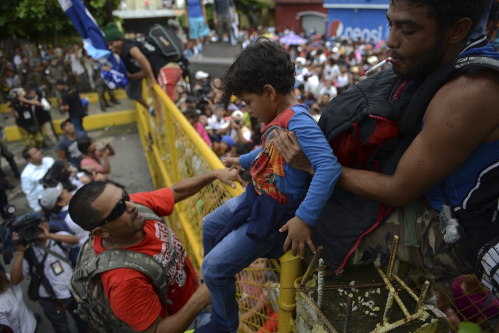 APTOPIX Central America Migrant Caravan A child is carried over the border fence as thousands of Honduran migrants rush across the border towards Mexico, in Tecun Uman, Guatemala, Friday, Oct. 19, 2018. Migrants broke down the gates at the border crossing and began streaming toward a bridge into Mexico. After arriving at the tall, yellow metal fence some clambered atop it and on U.S.-donated military jeeps. Young men began violently tugging on the barrier and finally succeeded in tearing it down. (AP Photo/Oliver de Ros)