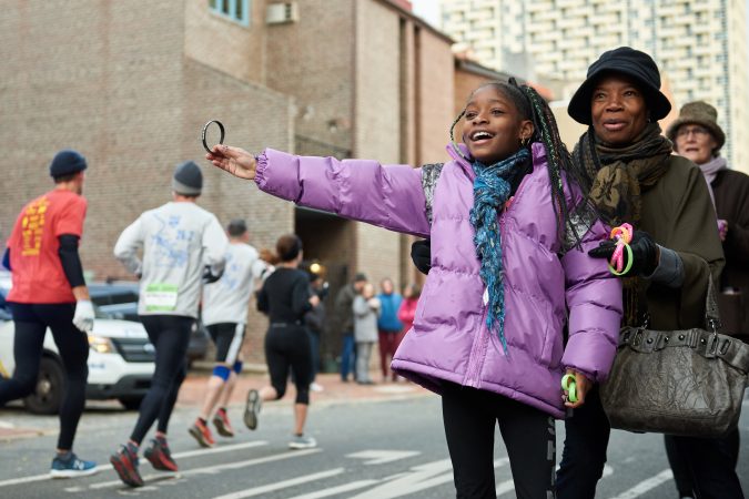 Brehana Mejia, 9, a young member of Mother Bethel A.M.E. Church, stands with Yvonne Hylton, and hands wristbands out to participants of the Philadelphia Marathon as they run past the church on Sunday, Nov. 18, 2018. She says her favorite part of the church is the singing. (Natalie Piserchio for WHYY)