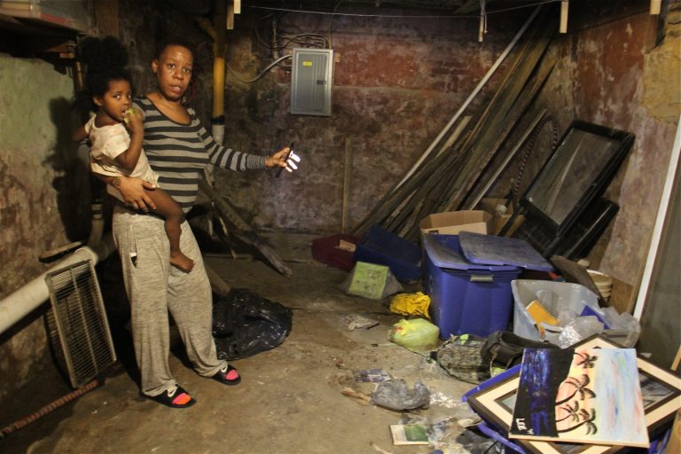 Sherri Lee, holding her 2-year-old daughter, Aubrey Boyd, shines a light on possessions that were destroyed when a backed up toilet flooded her basement. (Emma Lee/WHYY)