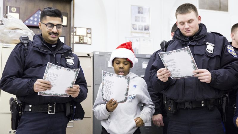 Officers Rana Ashfaq (left) and Joe Crouse flank Kofi Walker, 9, at the Caroling with a Cop event held in collaboration between the 39th Police District and the Georgia E. Gregory Interdenominational School Of Music on December 7. (Rachel Wisniewski for WHYY)