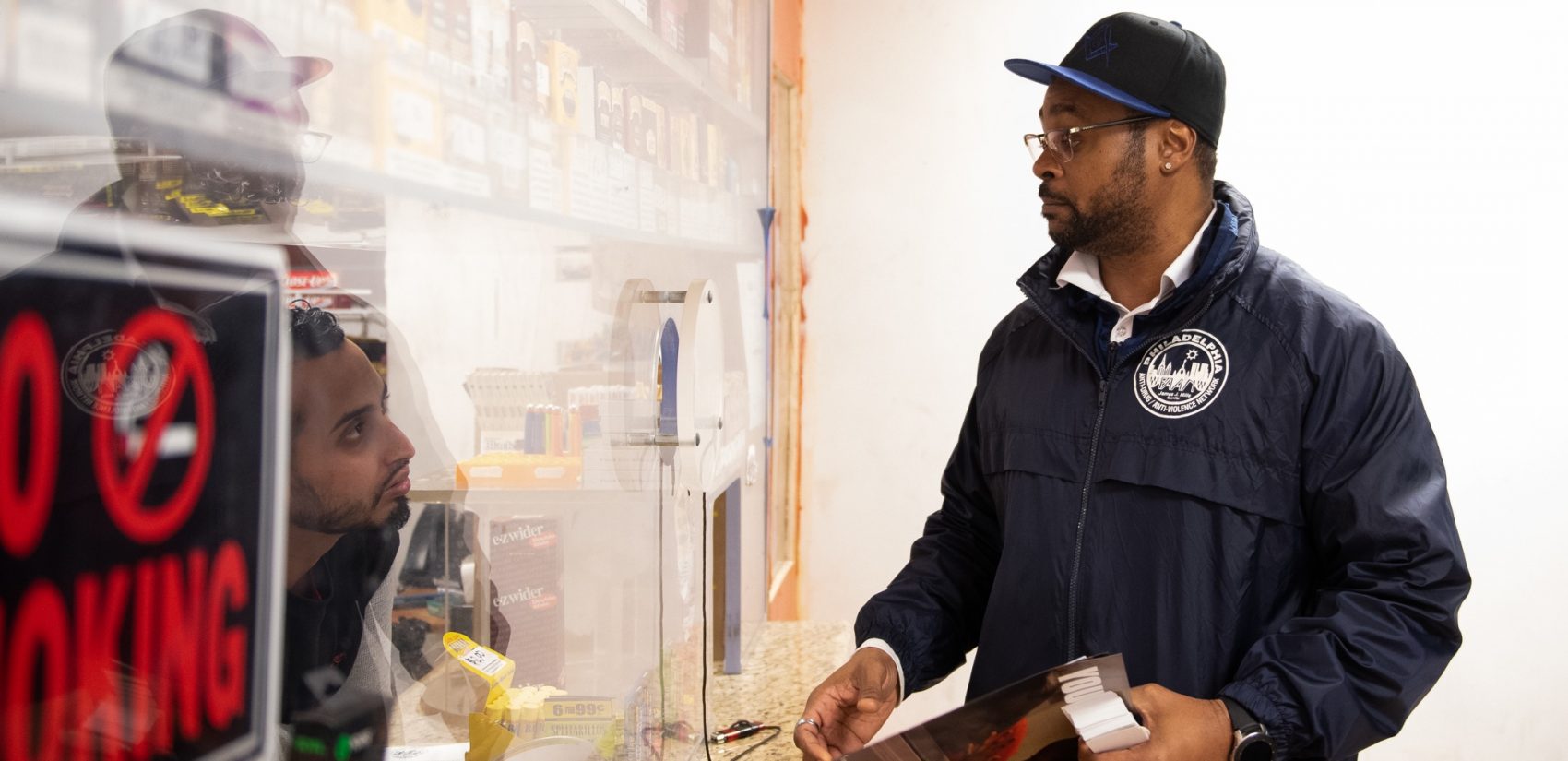 Tarik Harris talks to Sam, who's working the counter at the Logan Food Mini Market, about how CCIP can help businesses who are experiencing concerns of violence. (Kriston Jae Bethel for WHYY)