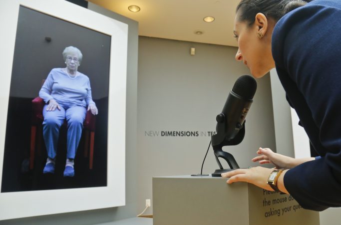 In this Friday Sept. 15, 2017, photo, Josephine Mairzadeh, (right), use a microphone to pose a question to a virtual presentation of Holocaust survivor Eva Schloss, (left), featured in a testimonial interactive installation called 