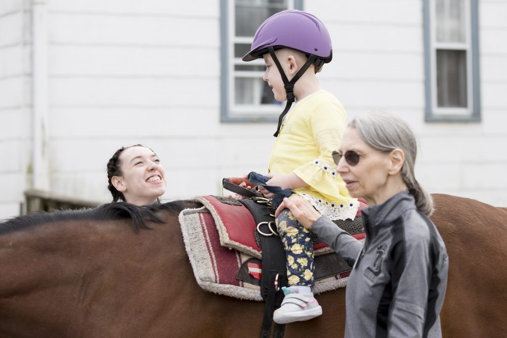 Volunteers Gabby Fink (left) and Carol Rowehl (right) help Emma Reed, 6, to stay upright on horse Prince Caspian on April 7, 2019. Reed is a participant in Pegasus Therapeutic Riding Academy's new Stepping Forward program for children of first responders. (Rachel Wisniewski for WHYY)