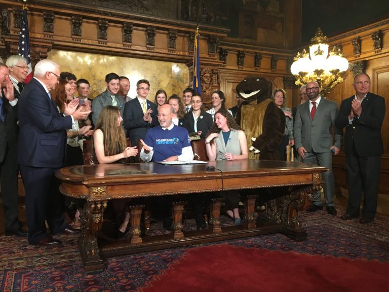 Surrounded by student supporters of the hellbender — and a mascot — Gov. Tom Wolf signs a bill officially designating the salamander as state amphibian. (Katie Meyer/WITF)