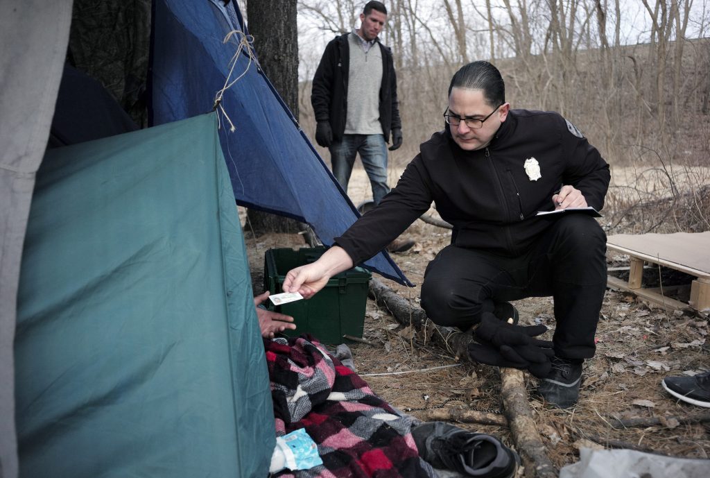 Hepatitis Outbreaks In this Tuesday, Feb. 12, 2019 photo Worcester Police officer Angel Rivera, right, returns a license to an unidentified man as Rivera asks if he has been tested for Hepatitis A at the entrance to a tent where the man spent the night in a wooded area, in Worcester, Mass. (Steven Senne/AP Photo)