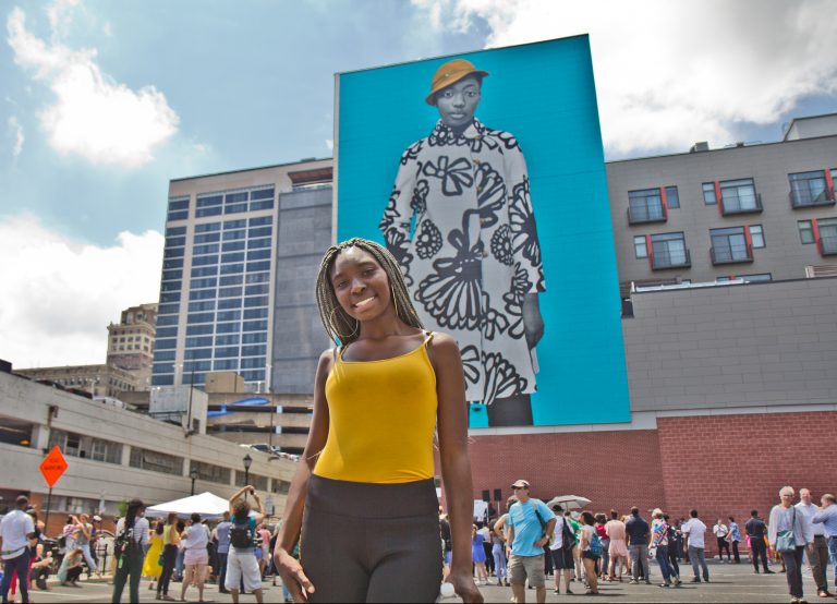 Najee Spencer-Young poses in front of her mural portrait by artist Amy Sherald. Spencer-Young is a participant in the Mural Arts’ Achieving Independence Center classes. (Kimberly Paynter/WHYY)