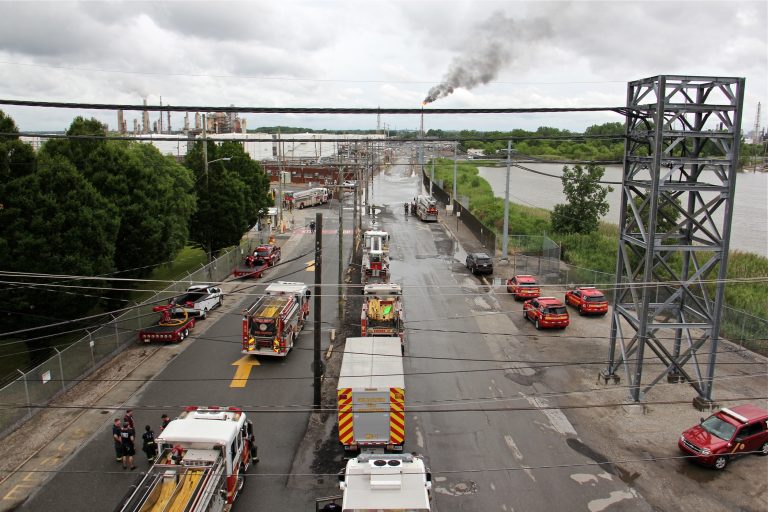 In this file photo, Philadelphia firefighters gather at the entrance to Philadelphia Energy Solutions Refinery, while a large flare burns off fuel to prevent it from feeding the massive fire at the refinery, Friday, June 21, 2019. (Emma Lee/WHYY, file)