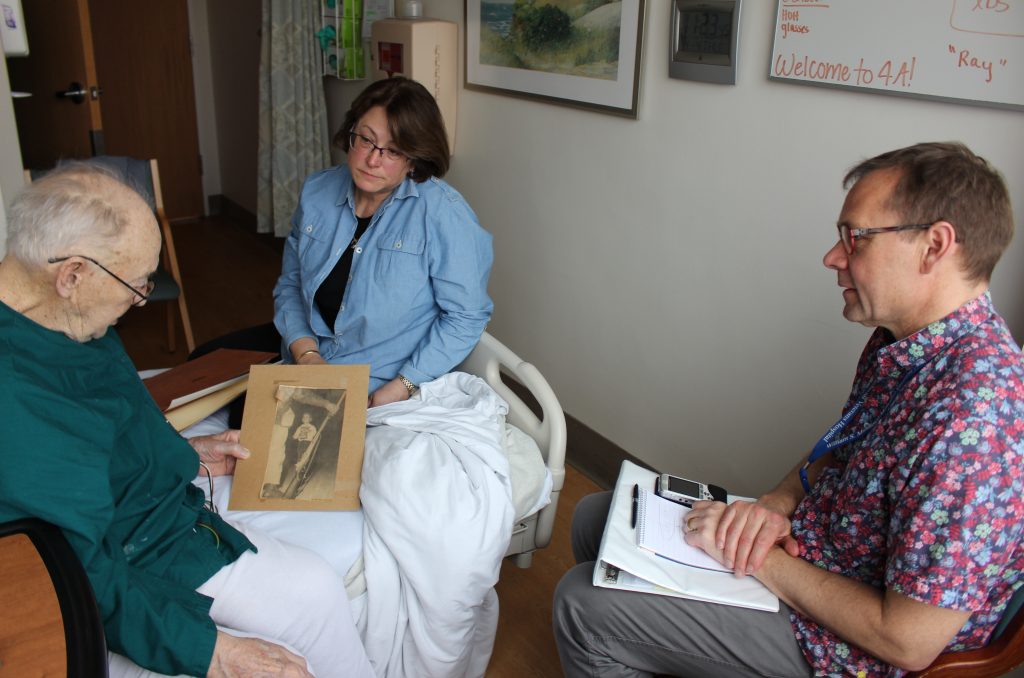 Thor Ringler (right) interviewed Ray Miller (left) in Miller’s hospital room at the William S. Middleton Memorial Veterans Hospital in Madison, Wis., in April. Miller’s daughter Barbara (center) brought in photos and a press clipping from Miller’s time in the National Guard to help facilitate the conversation. Thor Ringler (right) interviewed Ray Miller (left) in Miller's hospital room at the William S. Middleton Memorial Veterans Hospital in Madison, Wis., in April. Miller's daughter Barbara (center) brought in photos and a press clipping from Miller's time in the National Guard to help facilitate the conversation. (Bram Sable-Smith for NPR)