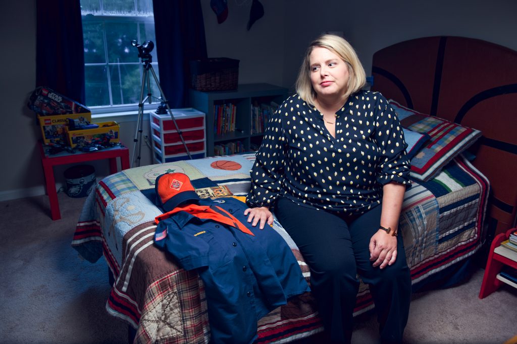 2019 08 14 boyscout bethel Leslie Slingsby, executive director of Mission Kids, sits on her sons bed with his Boy Scouts of America uniform and handbook at her Collegeville, Pa. home on August 14, 2019. Slingsby's nonprofit works on behalf of Montgomery County to investigate claims of child abuse and advocates for child protection around the world. (Kriston Jae Bethel for WHYY)